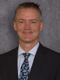 man looking and smiling at the camera wearing black suit and tie