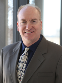 Photo of Dr. Goergen in a brown jacket with a black shirt and eye chart tie